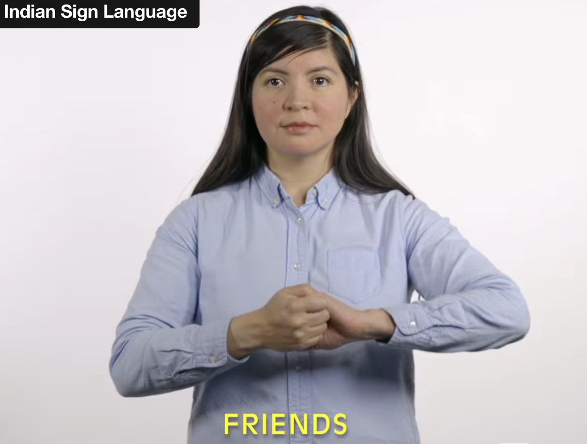 woman with long dark hair, light blouse, head band, with hands held together in front of her, caption in yellow on bottom center says FRIENDS, text at top left said INDIAN SIGN LANGUAGE white background